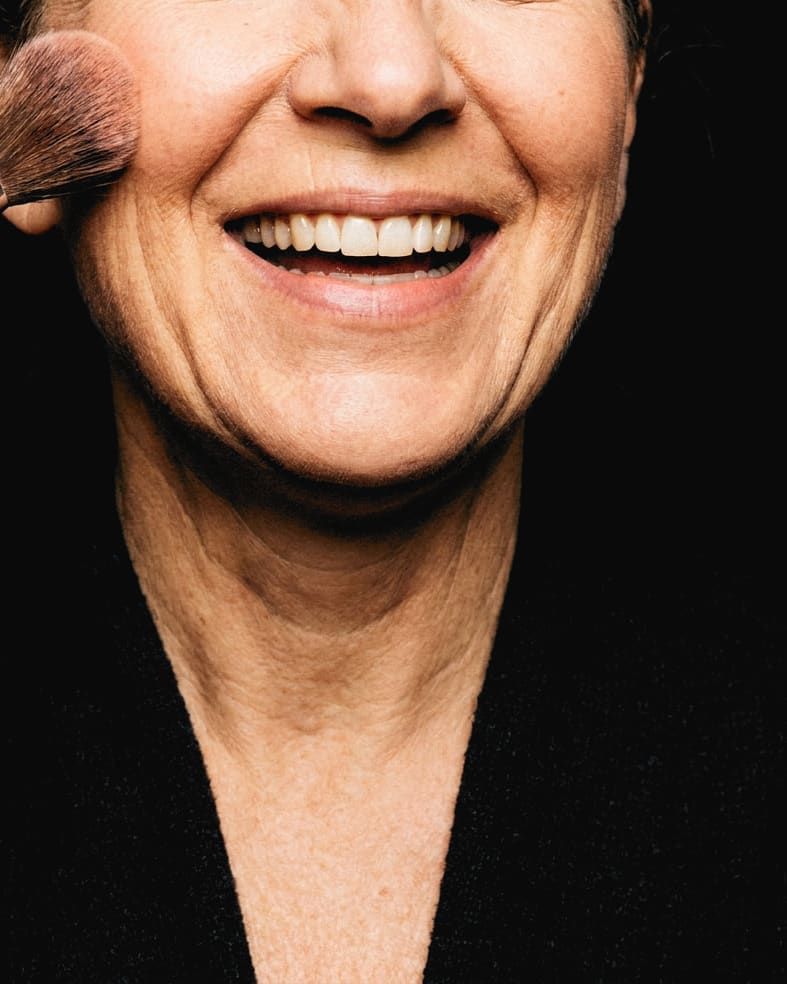 A blush brush sweeping a cheekbone, a woman mid laugh