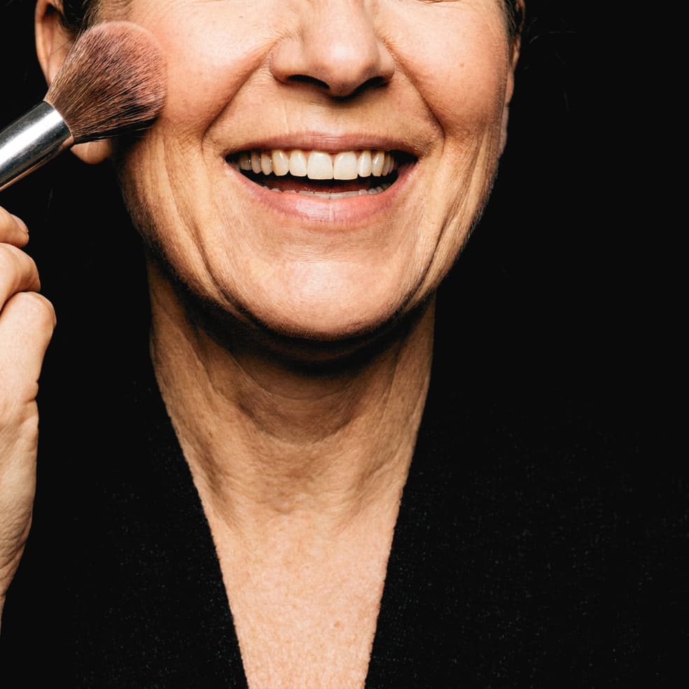 A blush brush sweeping a cheekbone, a woman mid laugh
