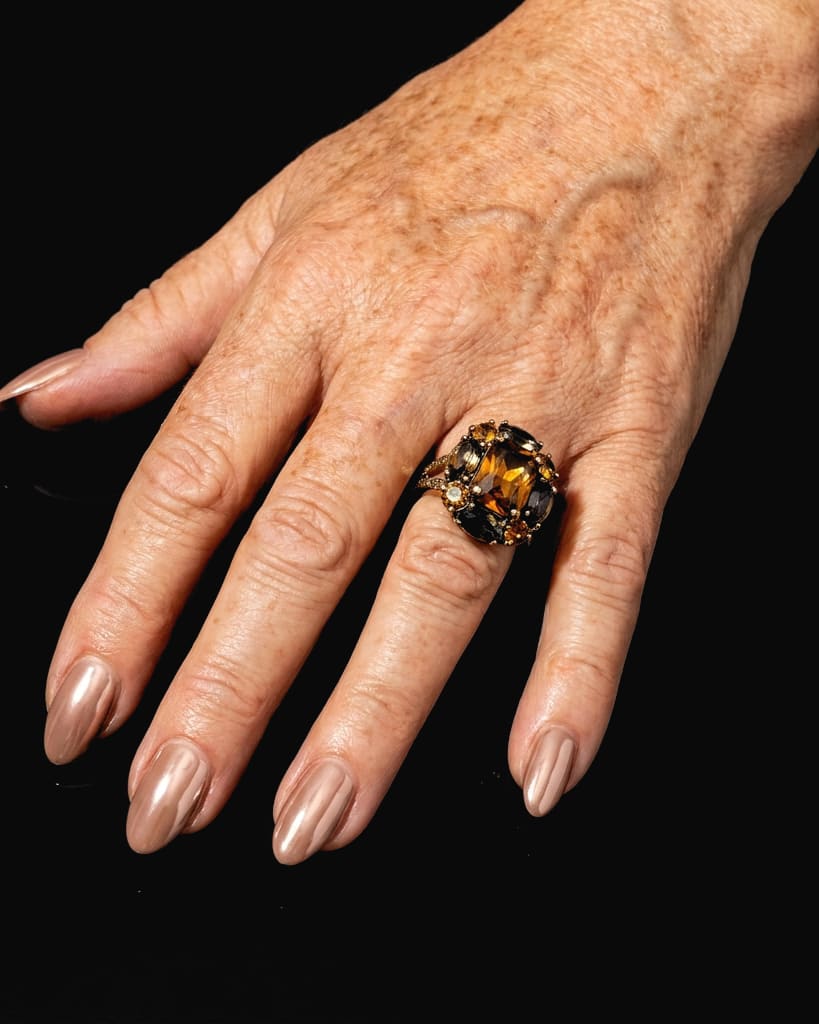 A freckled hand with glossy nude chrome almond nails and a big cocktail ring