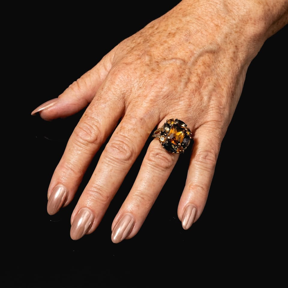 A freckled hand with glossy nude chrome almond nails and a big cocktail ring