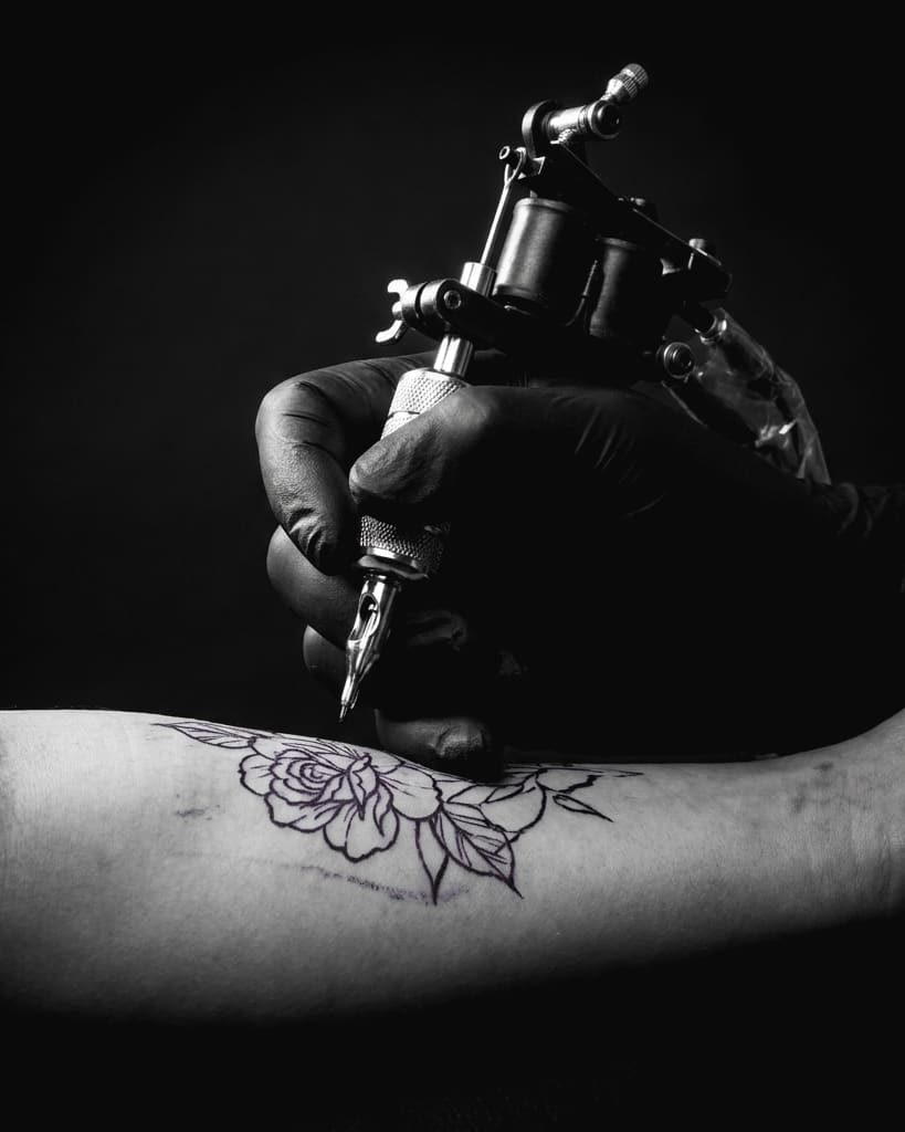A tattoo machine working over a rose stencil on a forearm, black and grey