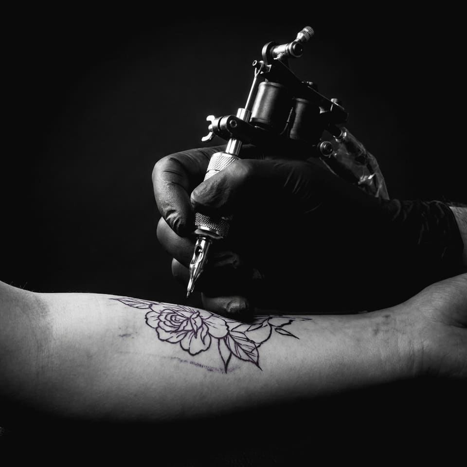A tattoo machine working over a rose stencil on a forearm, black and grey